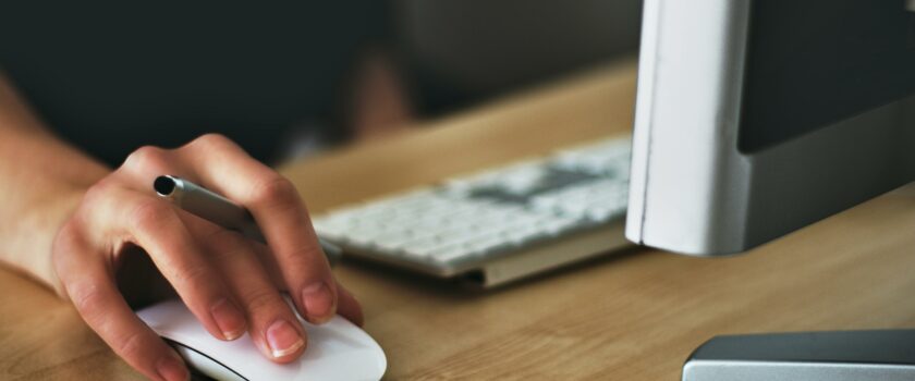 A hand using a wireless mouse at a modern desk setup with a computer and keyboard.