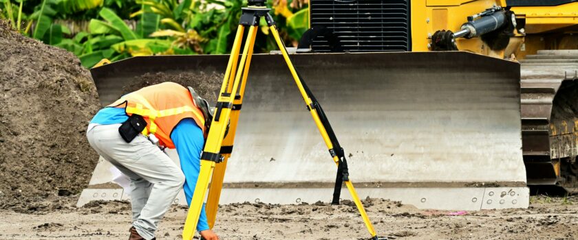 A construction worker operates a total station in front of a bulldozer on a construction site.